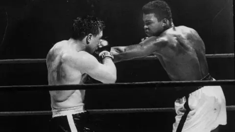 Getty Images Black and white photo of Muhammad Ali and George Chuvalo boxing in a ring. Chuvalo, in dark shorts with a white stripe, has his arms up, blocking a punch thrown by Ali, who is in white shorts with a dark stripe. 