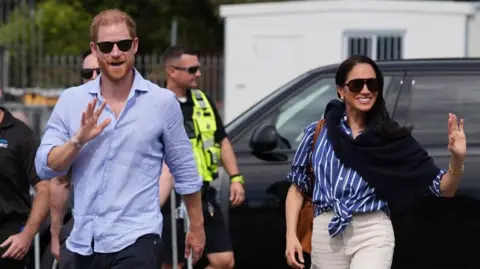 Reuters Harry and Meghan waving at a crowd, several security staff and a black car are behind them on 17 April 2026