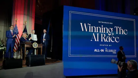 Getty Images US president Trump, flanked by several members of his administration, sits behind a desk with the presidential seal on stage at an event in 2025. To the right of him is a huge blue sign that says "Winning the AI Race" in white font. He is holding a folder containing an executive order he had just signed on AI.