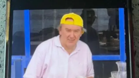 Supplied Image shows a man in a white/pale pink shirt, opened at the collar. He is wearing a yellow baseball cap, back to front, and some grey hair is sprouting out the sides. He looks like he may be on holiday. His eyes are squinting towards the camera and he has a slight smile. 