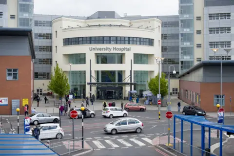 The exterior of University Hospital in Coventry. It has a semi circular entrance with windows and a wide door, with red brick buildings either side. Cars are parked outside the entrance