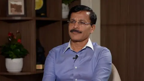 BBC Tukaram Mundhe wearing a blue shirt and black glasses looks away from the camera 