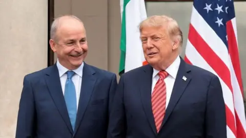 Reuters Micheál Martin to the left with blue suit and blue tie. Donald Trump to his right with dark suit and red tie. The Irish and American flag are behind them.