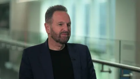 BBC Rene Haas sitting being interviewed. He has short brown hair and a short beard, and is wearing a dark blue blazer over a black T-shirt.