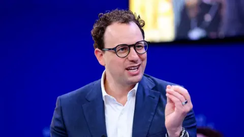 Reuters Dario Amodei hld the closed fingers of his left hand together as he mades a point. He is wearing a white shirt and blue jacket, black-rimmed glasses and has slightly curly hair. He is sat against a blue background, with a yellow-lit square just behind and to the left of his head. 