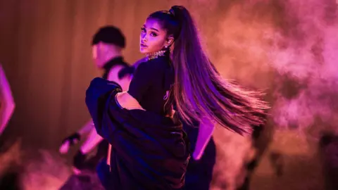 Getty Images Ariana Grande performing on stage. She has long hair in a ponytail. She is wearing a oversized black coat over a black t-shirt. 