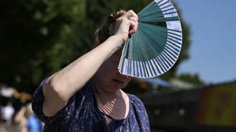 Getty Images A woman stands in the sun and holds a fan up to shade her face. She is wearing a short sleeve blue top with gold symbols. The background is blurred but the outlines of other people can be seen