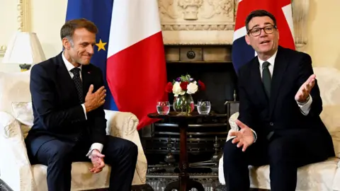 PA Wire Emmanuel Macron and Andy Burnham sit on chairs in Downing Street smiling and in conversation with their respective flags behind them.