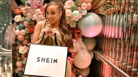 Getty Images for SHEIN A woman poses for a picture while holding a Shein-branded shopping bag. She is surrounded by pastel-shaded balloons in a room.