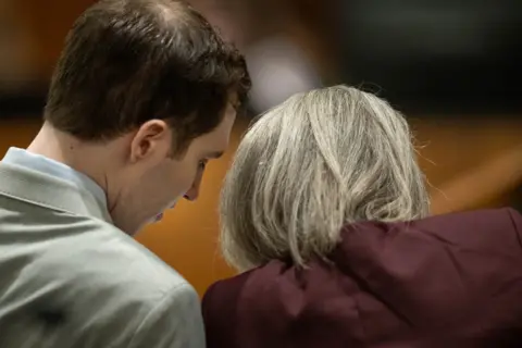 EPA/Shutterstock Tyler Robinson, wearing a grey suit and light-coloured shirt, leans in to consult with defence attorney Staci Visser, her blond-grey hair styled neatly to her shoulders over a maroon blazer; only the side of Robinson's face and the back of Visser's head can be seen