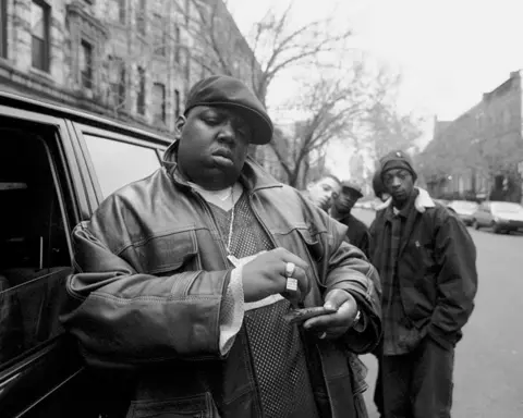 Getty Images In this black and white photograph taken in Brooklyn outside his mother's house, Wallace is in the foreground wearing a big leather jacket and a beret. He is standing next to a car and rolling a cigar while his friends stand in a group behind him. On the right you can see down what looks like a New York street