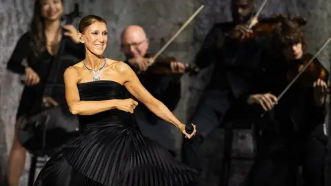 Getty Images Dion dancing in a black dress, holding a microphone, while musicians play behind her