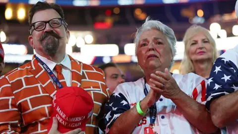 Getty Images A man in a brick wall suit stands during the convention. He is holding a Make America Great Again hat. Beside him, a woman stands clapping