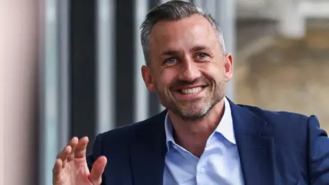 Reuters Ulrich Siegmund, who has greying hair and a short beard and wears a navy suit jacket with a light blue shirt, smiles and waves as he arrives at a press conference on Monday.