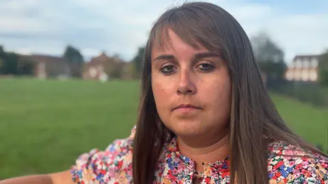 BBC Becky Jones has shoulder-length brown hair with a fringe. She is standing in a park and is wearing a dress with multi-coloured flowers on it. 