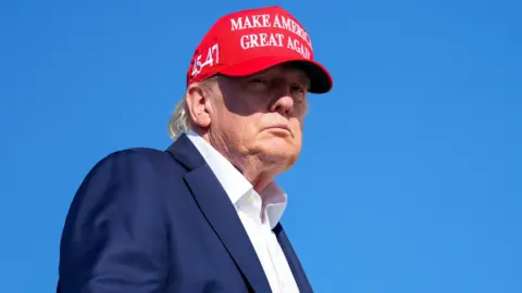 Reuters US President Donald Trump wears a blue suit jacket, white shirt and red cap, embazoned with the phrase MAKE AMERICA GREAT AGAIN in white writing. The sky is a deep blue behind him.