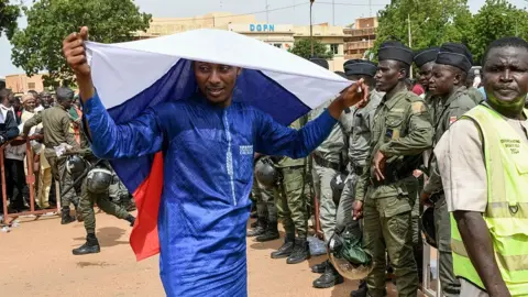 AFP via Getty Images A man in a blue boubou drapes a Russian flag over his head. A group of people in army uniforms can be seen to the right of the picture.