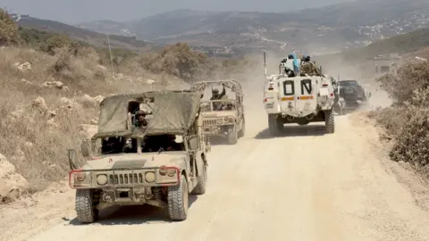 Neha Sharma/BBC Two Israeli military vehicles pass a Unifil convoy on a road along the wall separating Lebanon and Israel