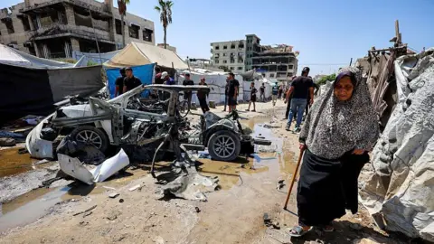 AFP via Getty Images An elderly woman walks past a destroyed vehicle following an Israeli strike in the Rimal neighbourhood of Gaza City (1 September 2026)