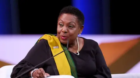 Getty Images Olivia Grange, a Jamaican woman with short black hair, is sitting down in front of a microphone. She is wearing black clothes and a black, yellow and green sash. She is wearing a pearl necklace and the background is blurred. 