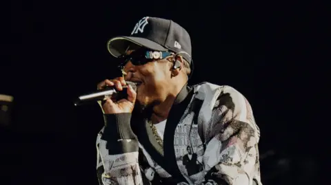 Roc Nation Jay-Z raps into a microphone while performing on stage. He wears a black and white cardigan and a New York Yankees hat.