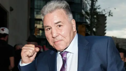 WireImage via Getty Images Close-up of Chuvalo, wearing a suit and tie, and holding up his fight and smiling