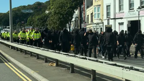 Lania Tkaczyk Dozens of men dressed in black and wearing face coverings, being escorted down a dual carriageway in Dover by uniformed police officers.