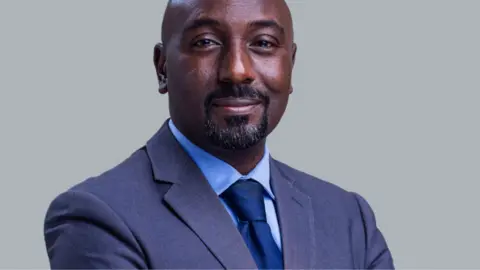 UBC Edward Rukidi Kijanangoma wearing a dark suit and tie smiling