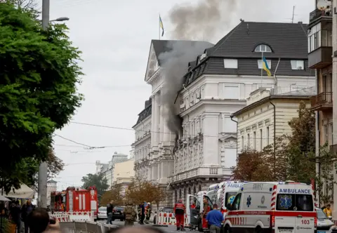 Reuters Black smoke pours from two windows in a large government building with Ukrainian flags on the facades. Ambulances and security personnel crowd the street below.