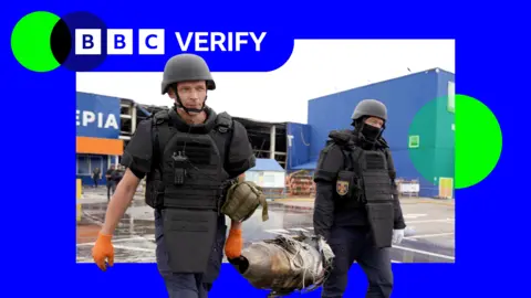 Getty Images Two Ukrainian first responders carry the remains of a Russian jet-powered drone engine after an attack. They are wearing body armour and helmets and are holding opposite ends of the engine. 