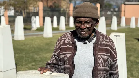 BBC Abraham Mofokeng in a brown patterned cardigan and beanie hat stands by a white monolith. Out of focus, in the background, are other white monoliths - they are part of a memorial.