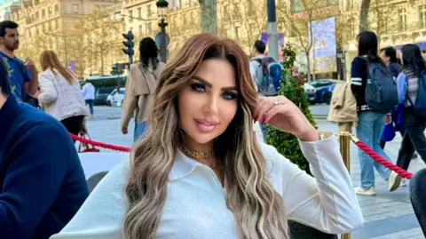 Sarah Khalifa Instagram account Sarah Khalifa wearing a white long-shirted with her folded left arm, the hand touching her long hair, sits by the side of a road inside a red-roped cordoned area, beyong which people walk on a road the other side of which has rows of sunbathed buildings