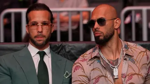 Reuters Tristan Tate is wearing sunglasses and is dressed in a green suit and tie and white shirt. Andrew is to his right in sunglasses and an open necked shirt.