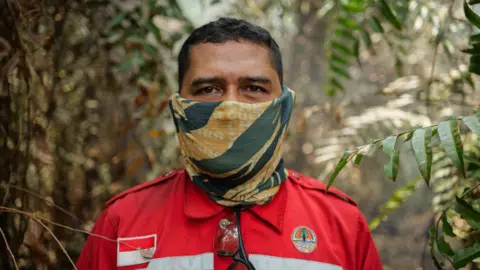 Ahmad Dhani for BBC A man in red firefighting uniform patched with Indonesia's flag, and with a yellow and green neck gaiter pulled over his face, stands in a forest and looks into the camera