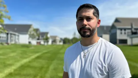 Supplied Gerald Bourguet, a white man standing in a field of a manicured lawn. He is wearing a white shirt and crucifix chain. There are homes in the background that are blurred.