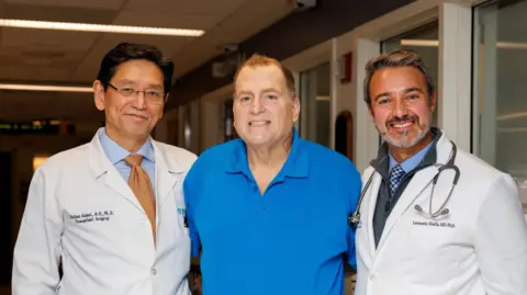 Kate Flock/Massachusetts General Hospital The image shows three adults standing closely together in a hospital hallway. The person in the centre is wearing a bright blue polo shirt, while the individuals on the left and right are wearing white medical coats, with a stethoscope visible around the neck of the person on the right. Fluorescent ceiling lights illuminate the corridor, and doors and windows line the walls behind the group. Small embroidered text is visible on the medical coats.