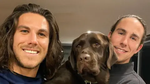 Instagram@Callum10Robinson A young man with shoulder-length brown hair and light stubble smiling at the camera, and another young man to his right and a brown dog in between them