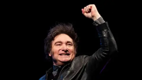 Tomas Cuesta/Getty Images Argentine President Javier Milei, wearing a leather jacket, stands smiling with a closed fist raised against a black backdrop.