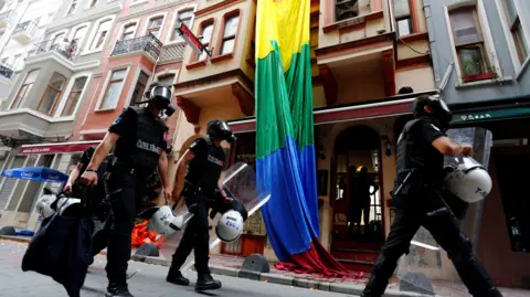 Reuters Riot police, with a rainbow flag in the background, chase LGBT activists as they try to gather for a pride parade, which was banned by the governorship in Istanbul. Officers are wearing black uniforms, wearing gas masks, and are carrying helmets and riot shields as they move through what appears to be a residential street. 