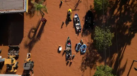 Florian Plaucheur/AFP via Getty Images An aerial shot showing boats moving through brown floodwaters in a street. To the right are some trees and telephone wires, to the left is a corrugated iron building and some machinery. 