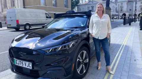 BBC Zoe Kleinman in blue jeans and cream blouse standing next to a dark Wayve self-driving robotaxi on a London pavement.