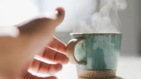 Getty Images A woman reaches her hand out to pick up a steaming hot mug which is shiny and green-coloured
