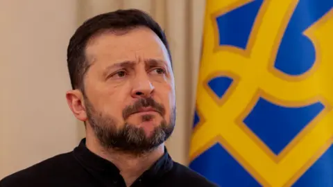 AFP via Getty Images Ukraine's President Volodymyr Zelensky gives a press conference in Kyiv, standing in front of a blue and yellow flag.